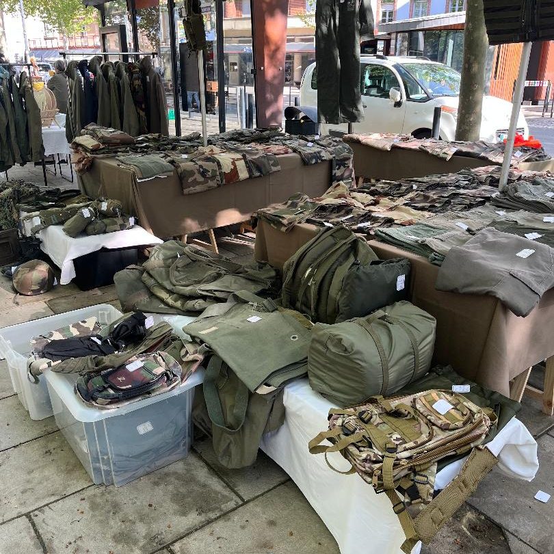 destock-and-compagny surplus de vêtements militaire Vêtements militaires occasion, treillis et veste kaki exposés sur des tables dans un marché en plein air.